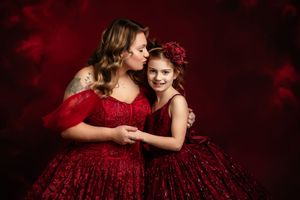 Two people in matching red sparkly gowns embrace against a dark red background in an elegant portrait setting.