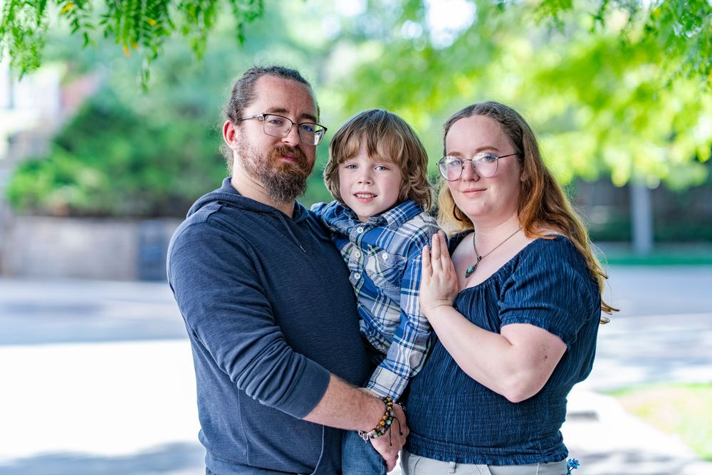 A young family of three poses outdoors, parents holding their toddler son among green trees.