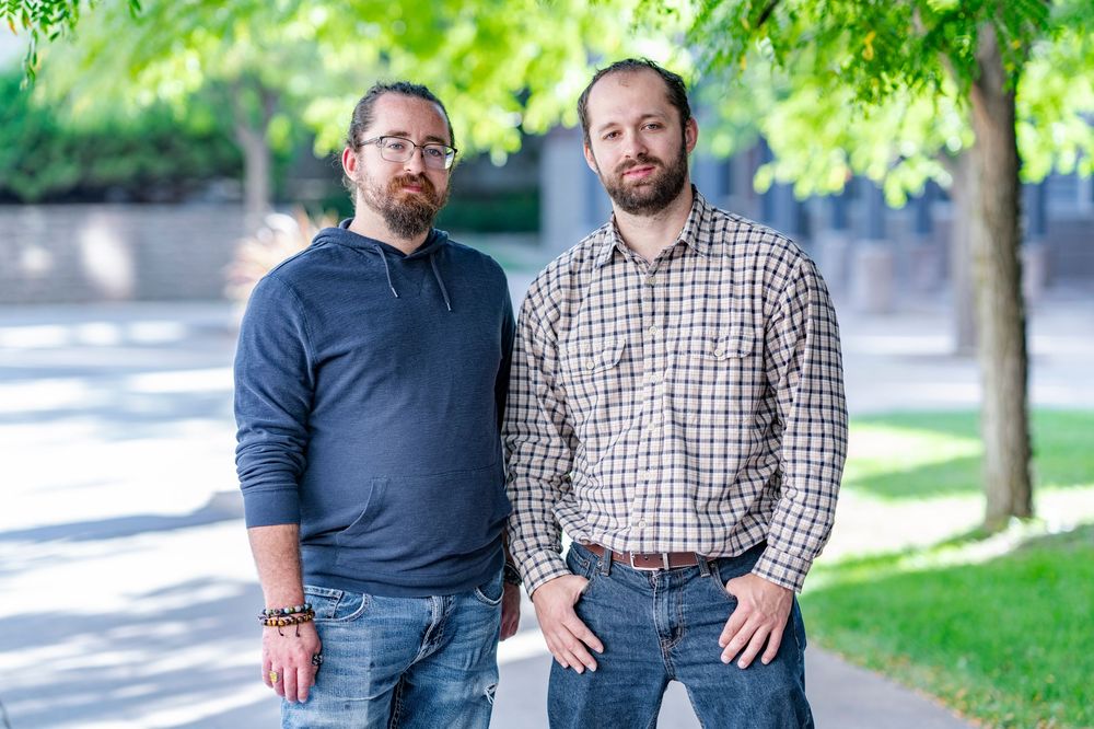 Two bearded men stand together outdoors under lush green trees on a sunny day.