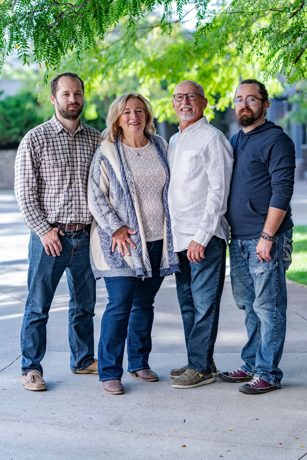 A family of four, two adult sons with their parents, pose together outdoors under green trees.