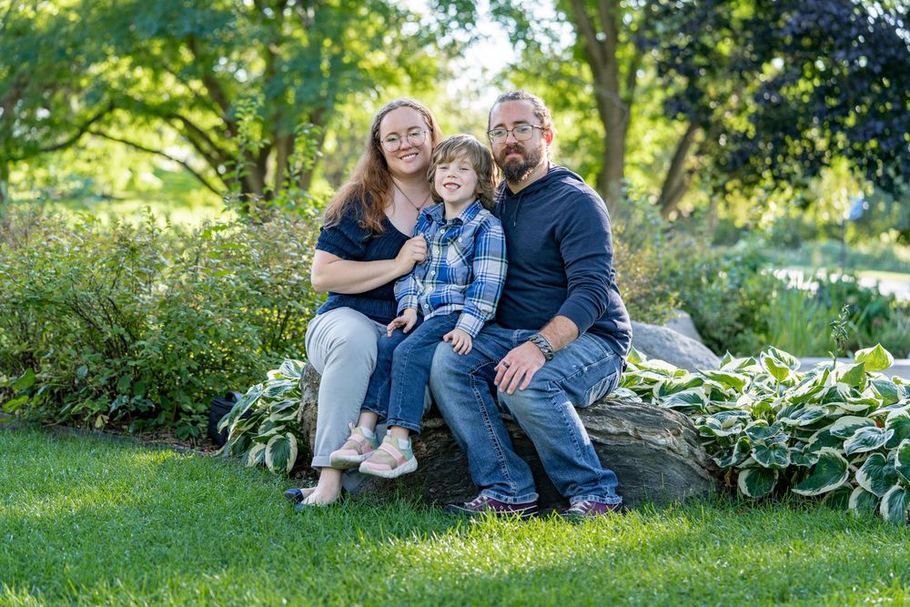A happy family of three sits on a stone in a lush garden, smiling at the camera.