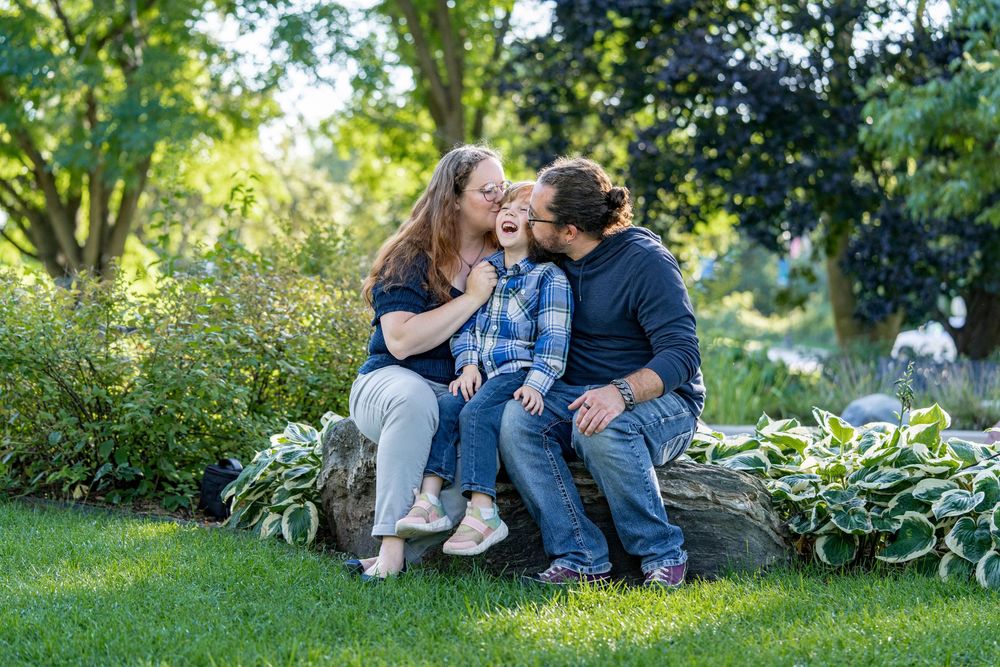 A joyful family of three shares a laugh while sitting on a stone surrounded by garden greenery.