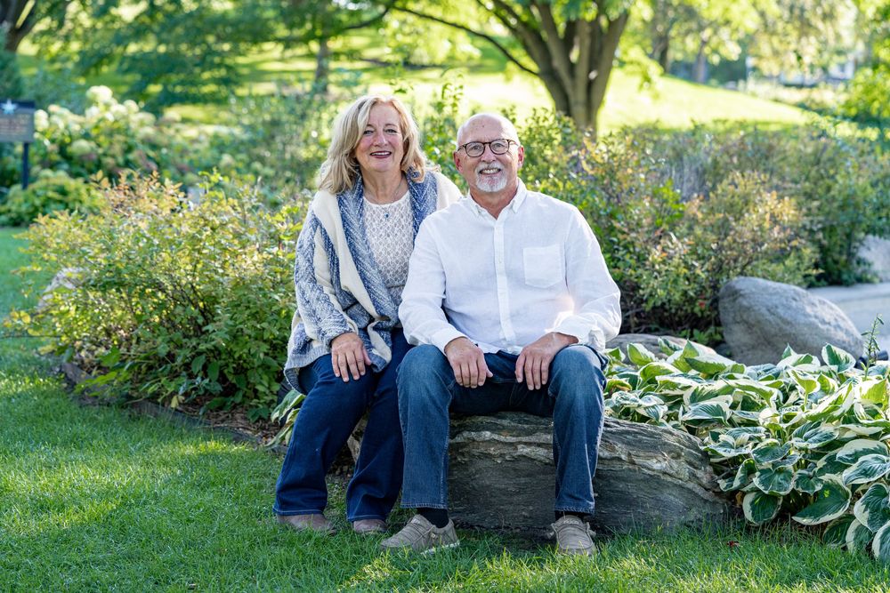 A smiling older couple sits together on a stone in a beautiful lush green garden.