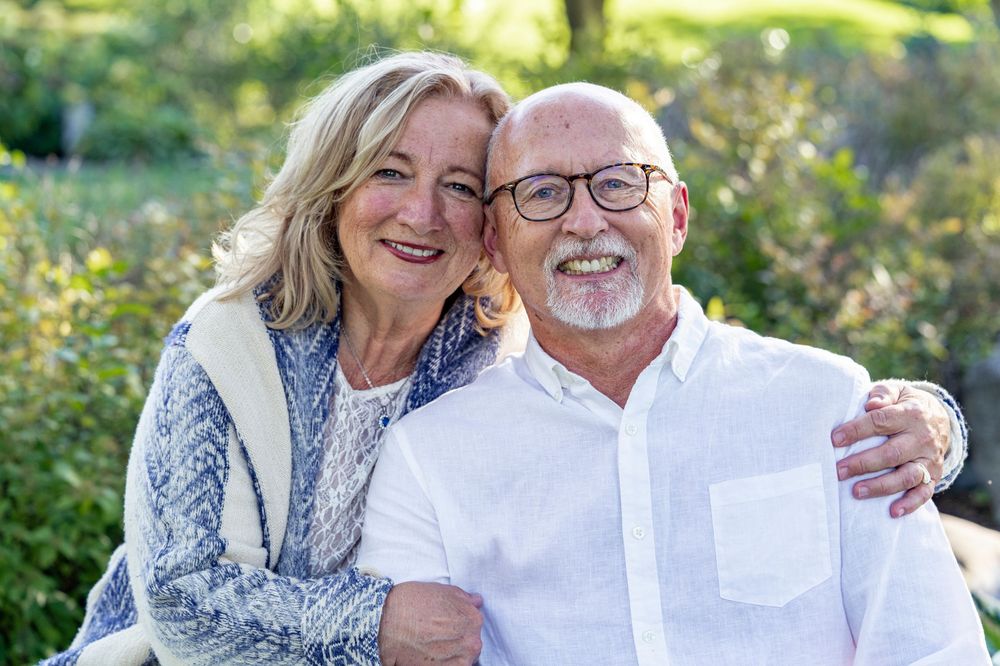 A happy older couple embraces and smiles warmly while sitting outdoors in a green garden.