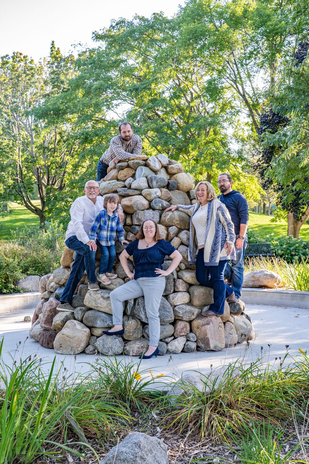 A multigenerational family of seven poses playfully on a large rock pile in a park.