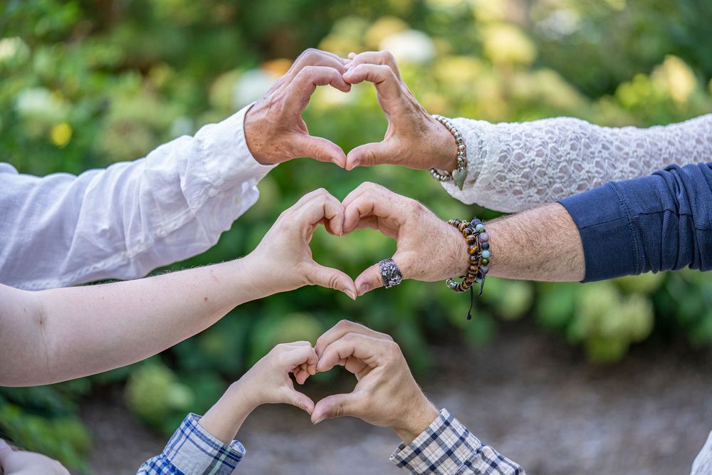 Three pairs of hands form heart shapes together against a soft green outdoor background.
