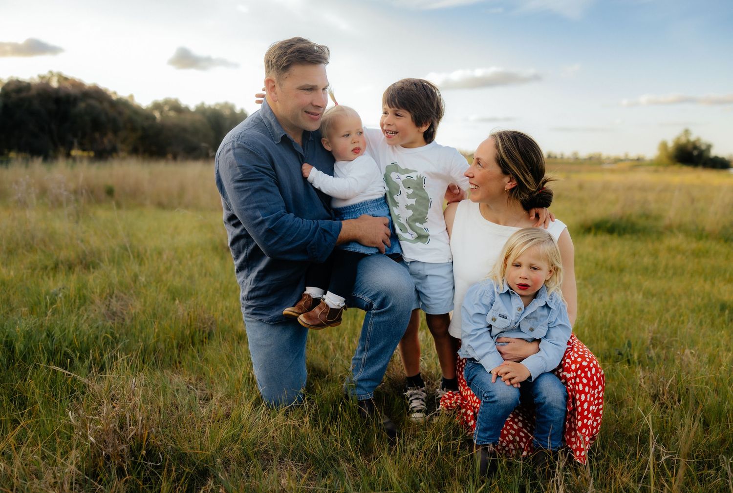 A joyful family of five shares a candid moment in a sunlit field, dressed in casual, coordinated outfits with warm smiles and laughter.