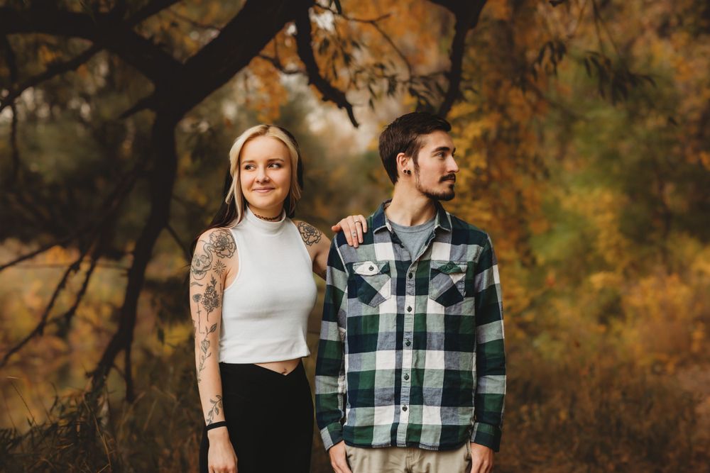 Two people stand together in an autumn forest setting with golden foliage and moody lighting in the background.