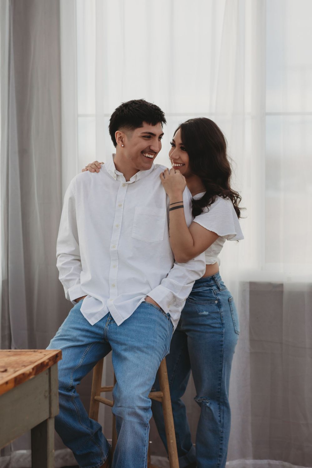 A couple wearing casual white and denim outfits share a candid laughing moment near a window with sheer curtains.