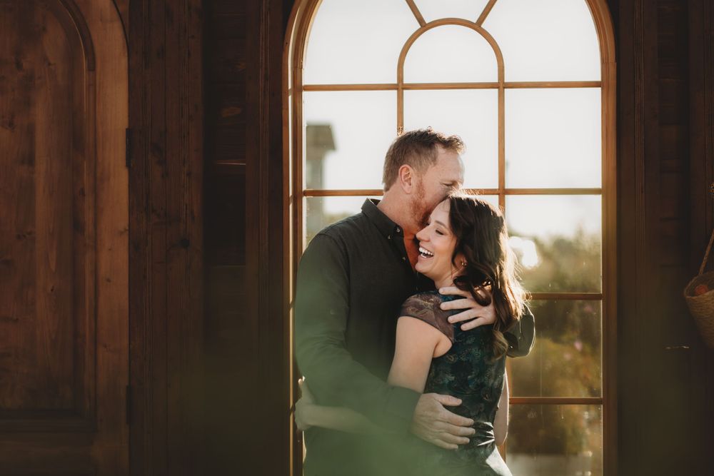 A romantic couple embraces by a sunlit arched window in a warm, intimate moment.