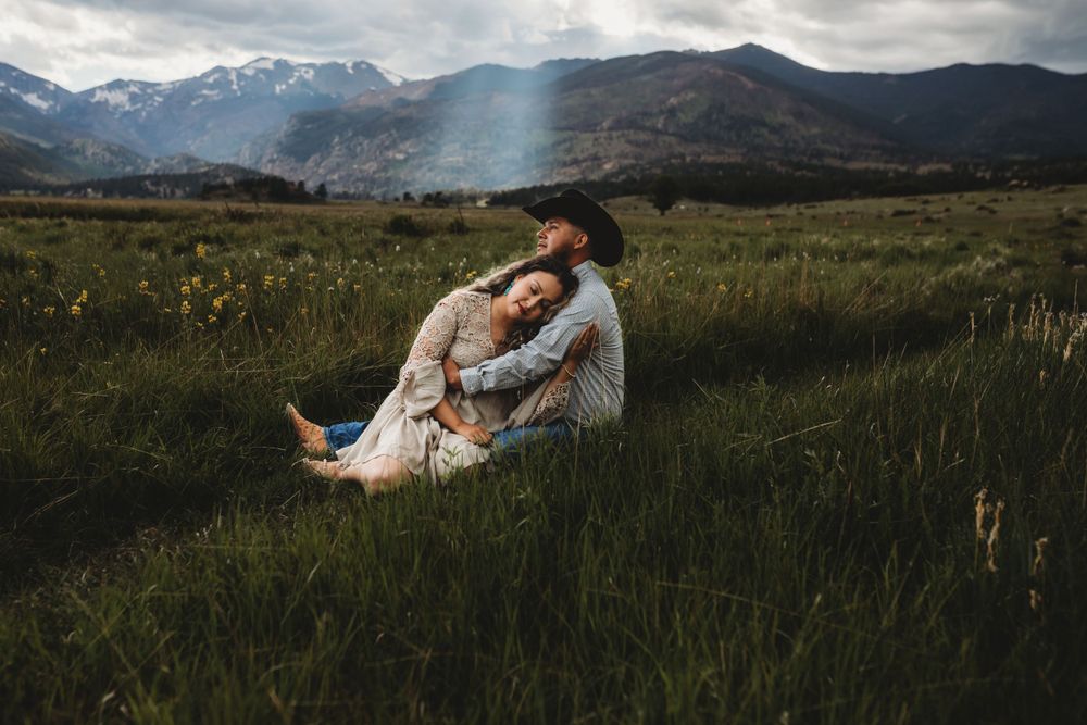A couple embraces while sitting in a mountain meadow with snow-capped peaks in the distance at sunset.