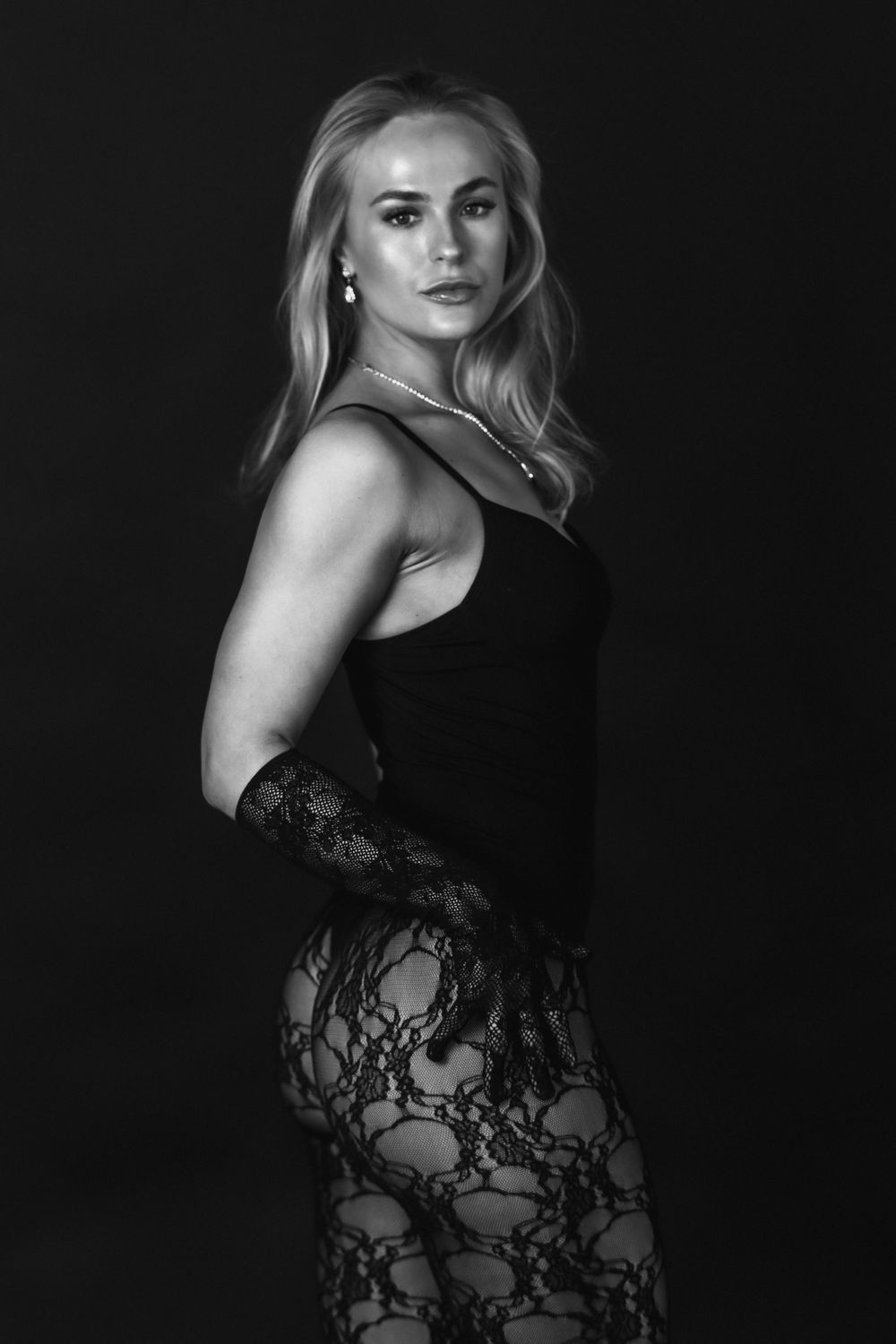 Black and white portrait of a blonde woman in a black bodysuit and lace gloves posing confidently.