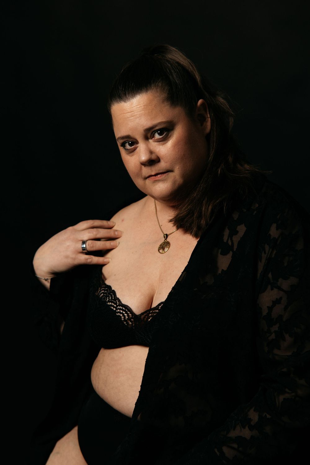 Plus-size woman in black lace lingerie gazing forward against dark background.