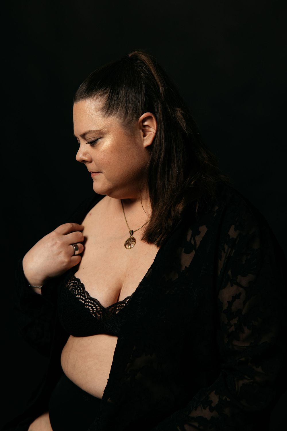 Plus-size woman in black lace bralette and jacket posing on dark background.
