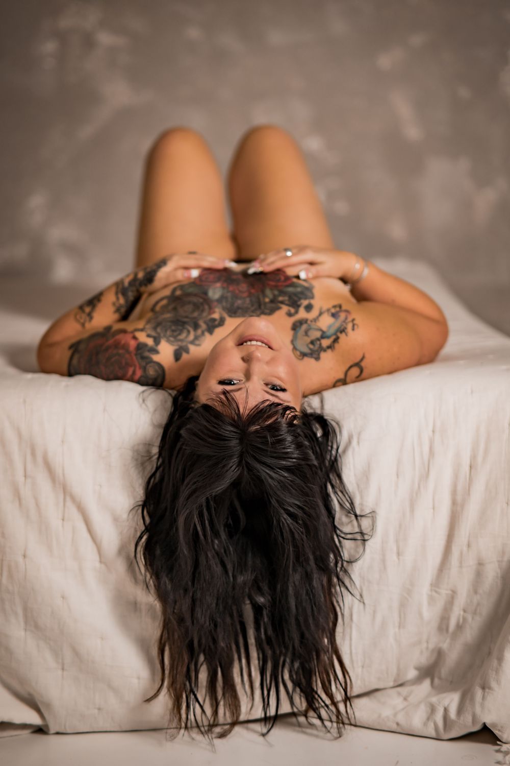 Tattooed woman lying on a bed with dark hair spread out, looking upward.