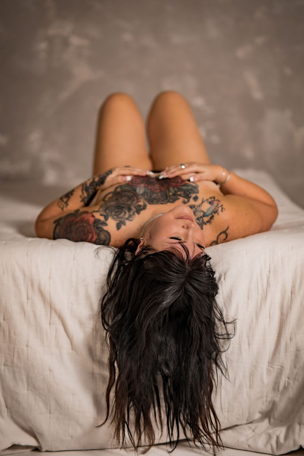 Heavily tattooed woman reclining on white bedding with knees raised.