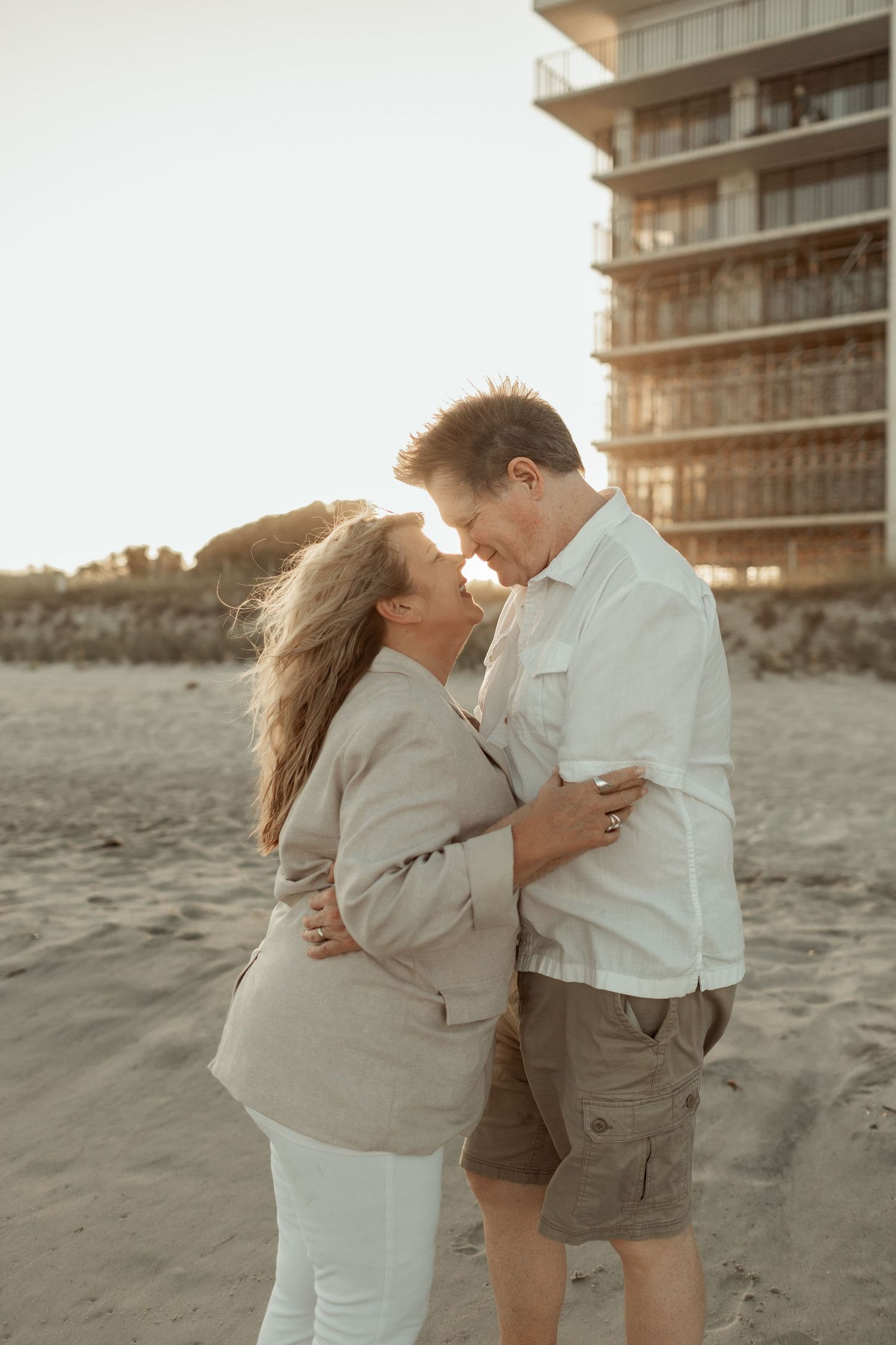 Romantic couple embracing on beach at sunset with apartment building in background.