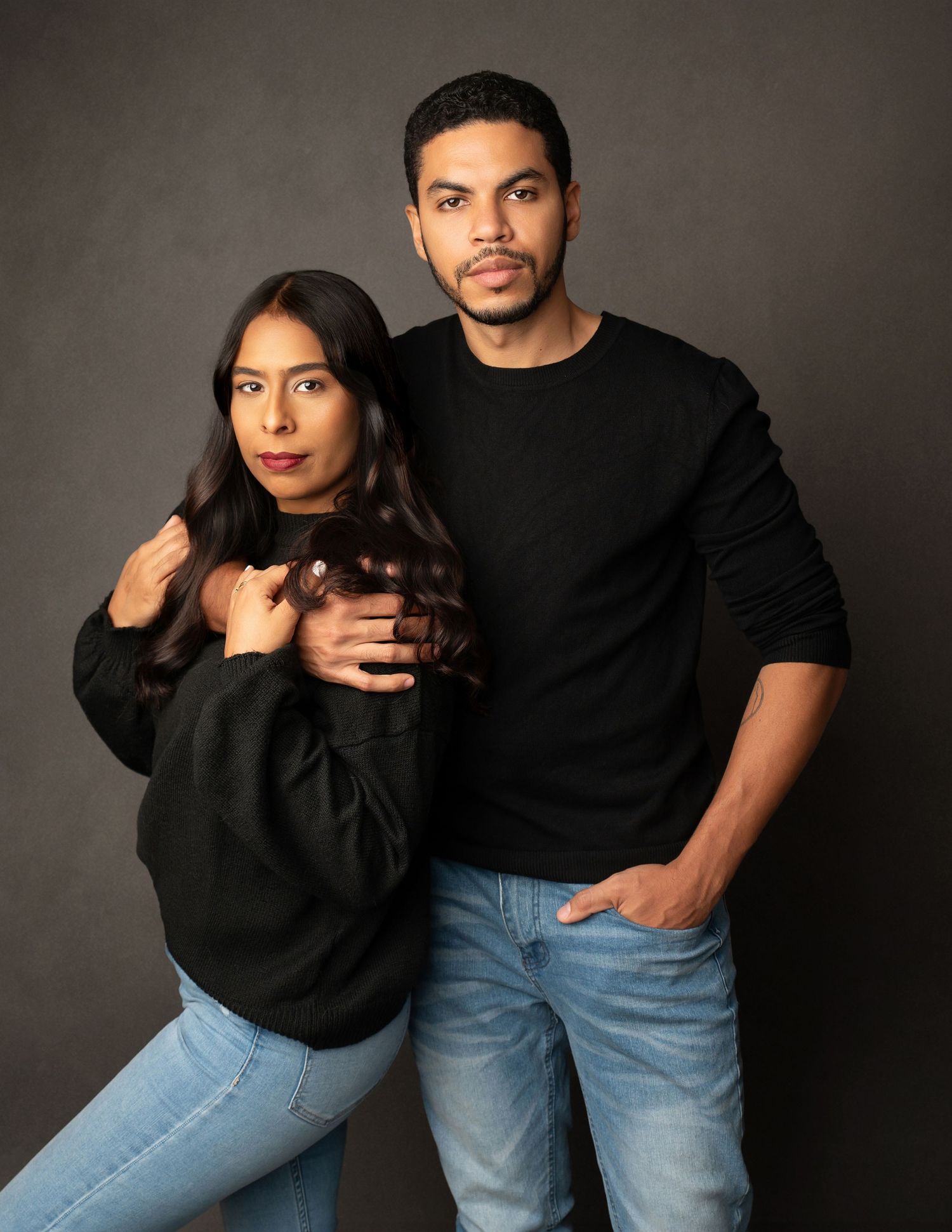 A couple wearing black sweaters pose together against a gray backdrop, creating an intimate studio portrait.