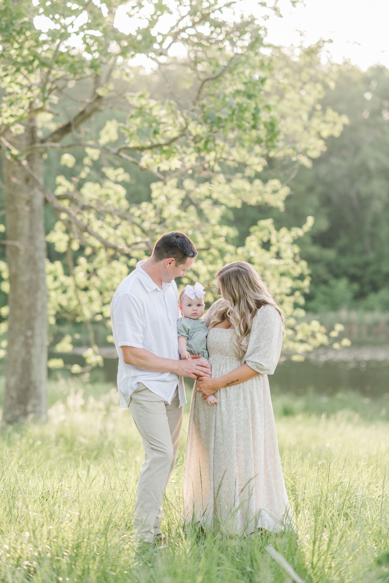 A young family with a baby stands in a sunlit meadow beneath a blossoming tree during golden hour.