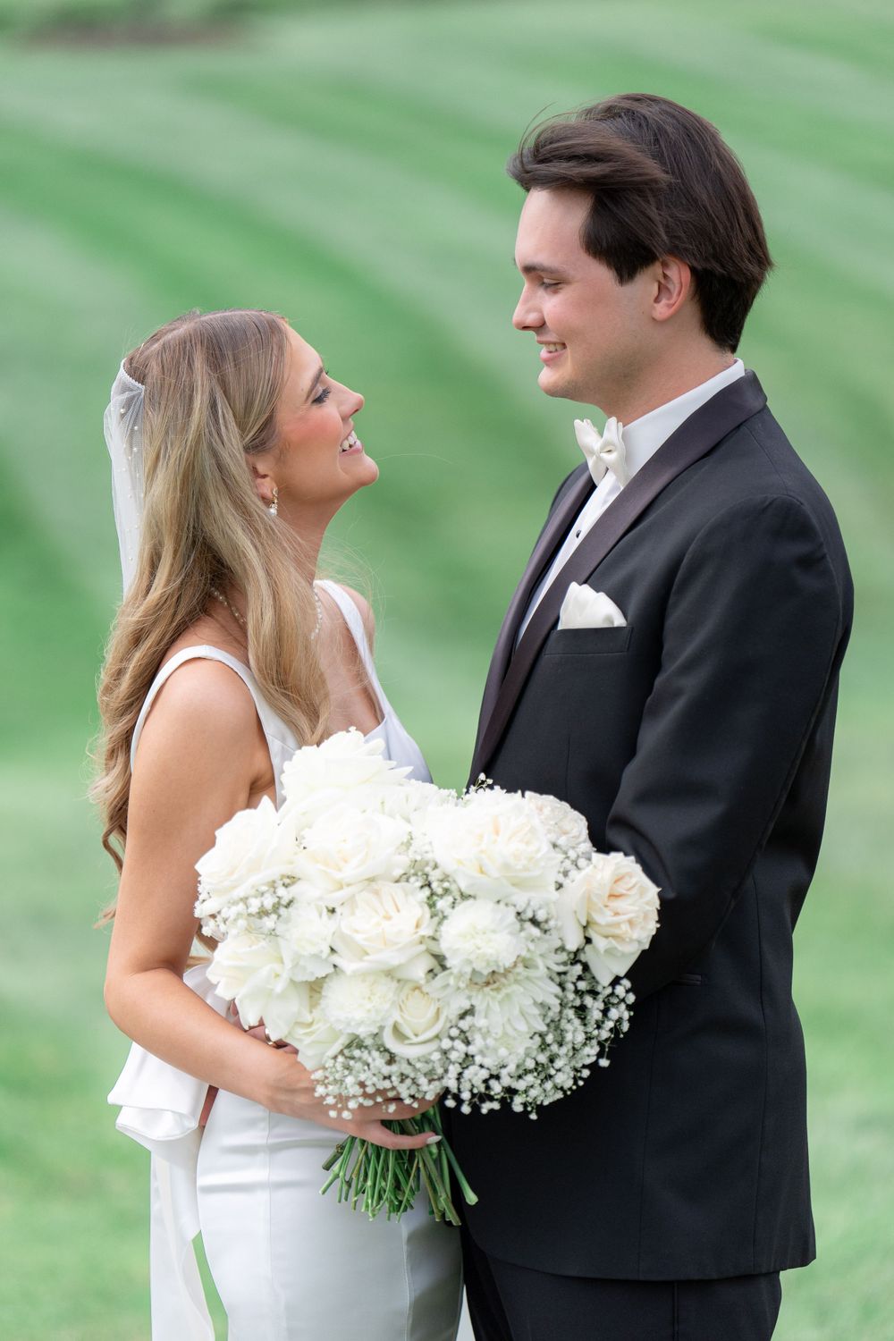 Bride holding white rose bouquet while sharing intimate moment with groom on grassy hill.