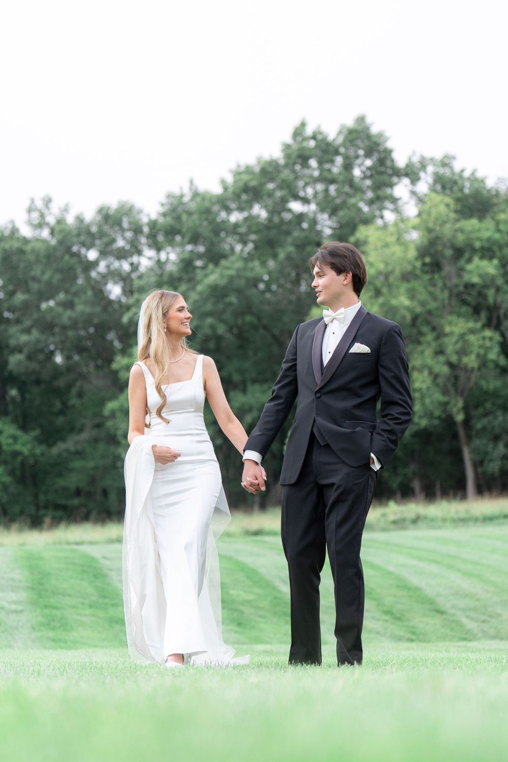 Newlyweds holding hands while walking on manicured lawn with trees in background.