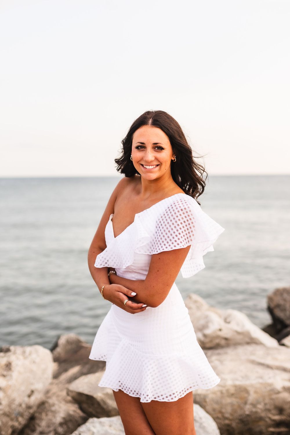 A senior girl in a white dress standing on the rocks on the shore of Lake Michigan.