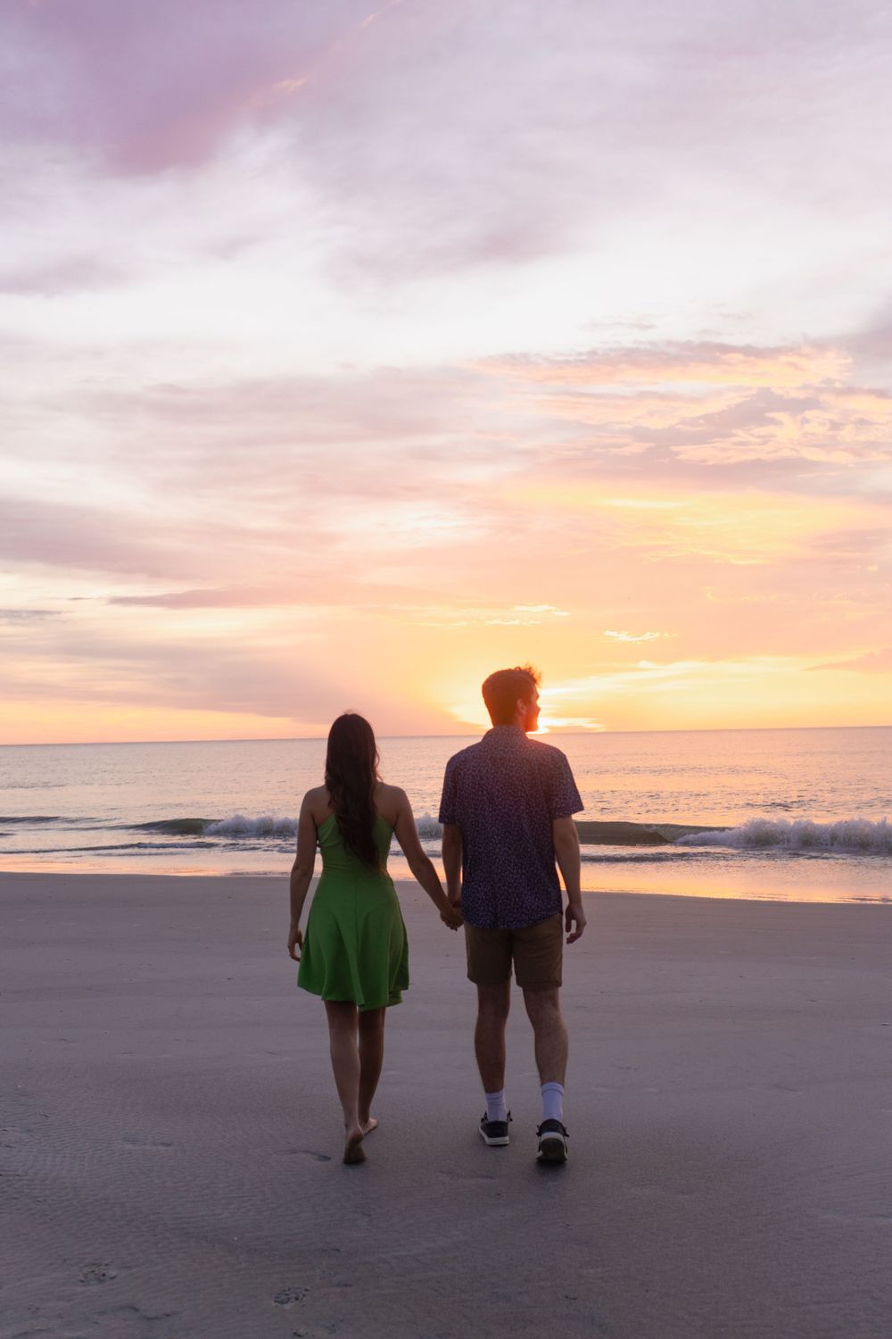 Two figures stroll along a peaceful beach at sunset casting silhouettes against the orange and pink sky.