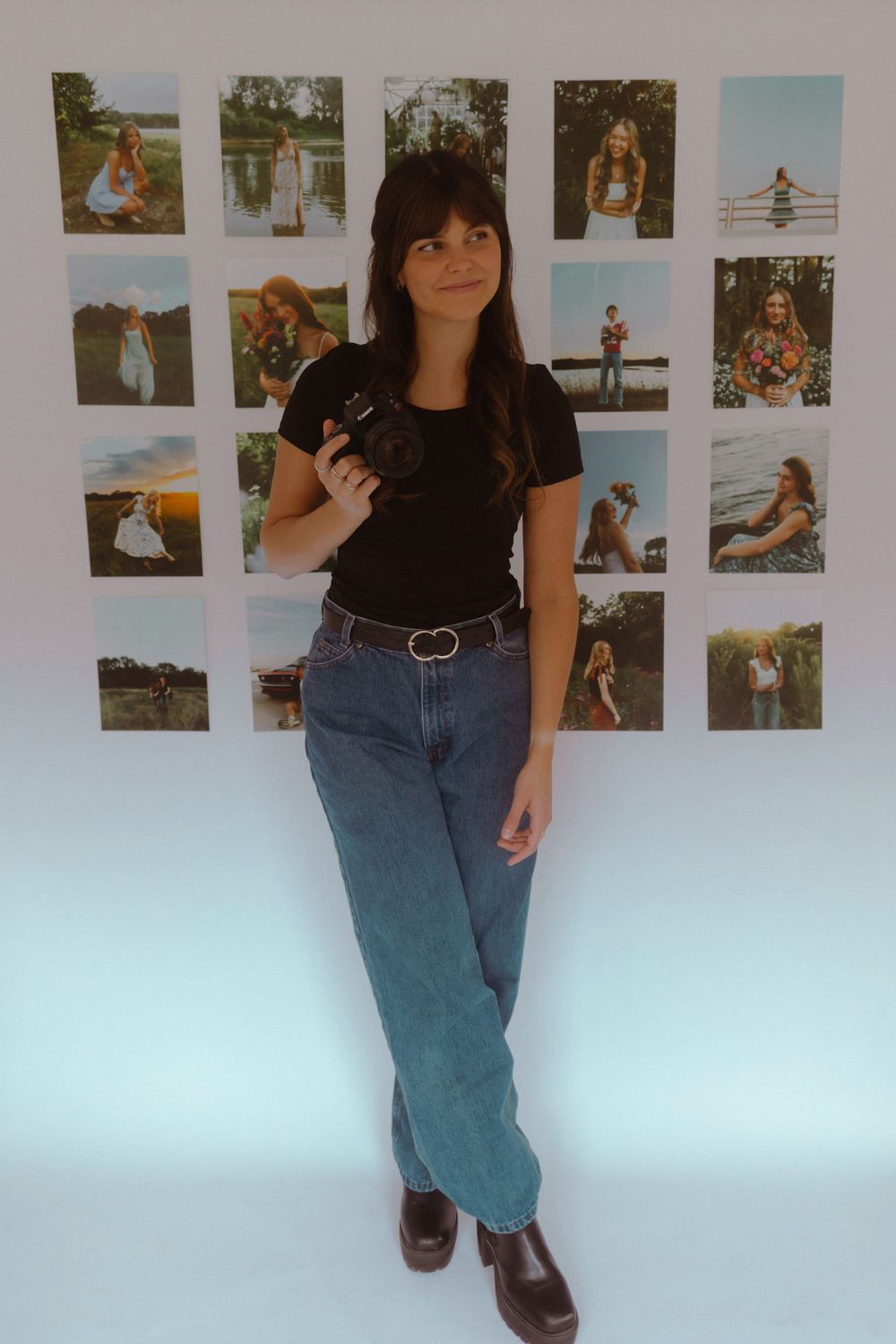 Person wearing a black top and blue jeans poses against a white wall decorated with casual photo prints.