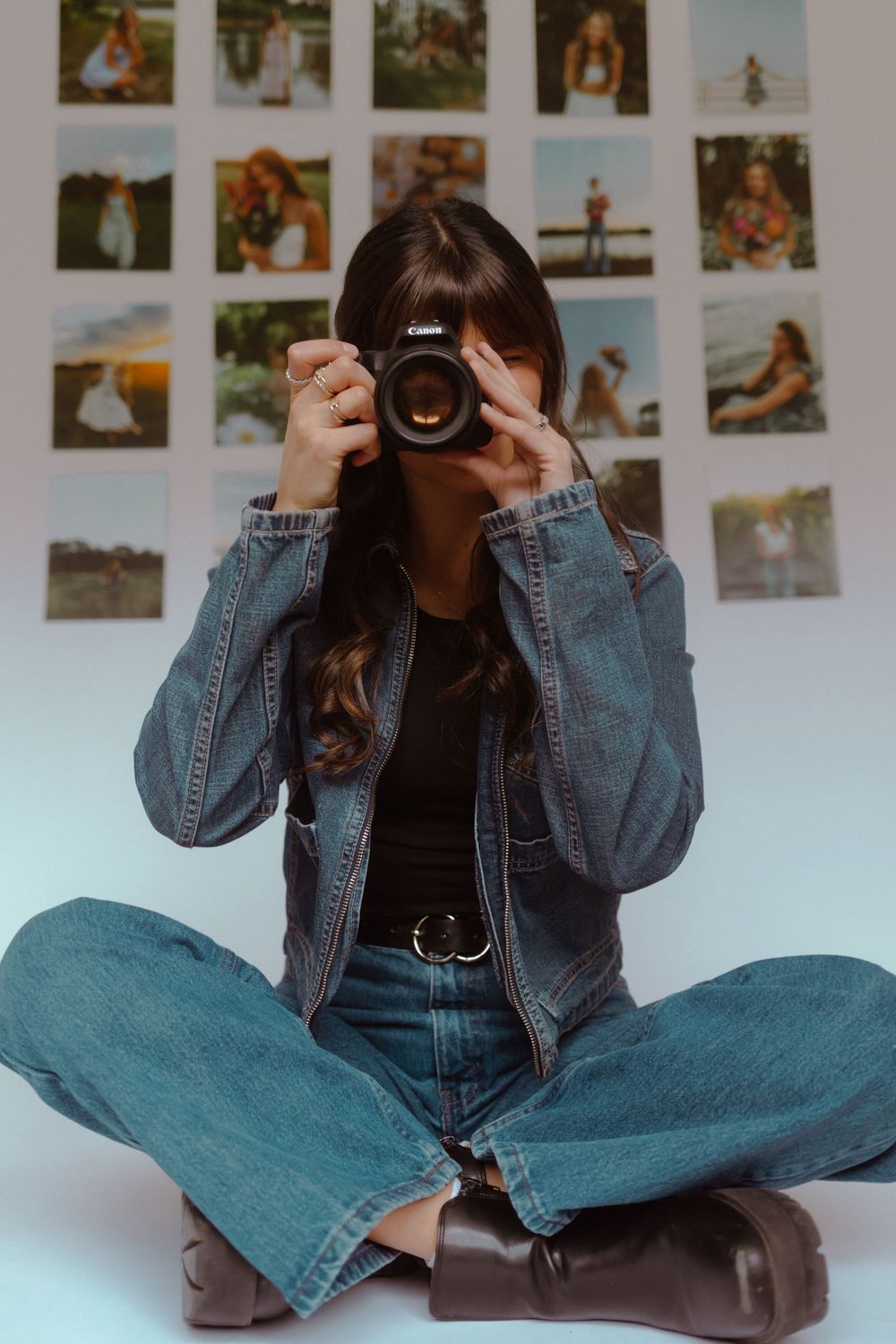 Person in denim jacket sitting cross-legged on floor holding camera against wall decorated with photographs.