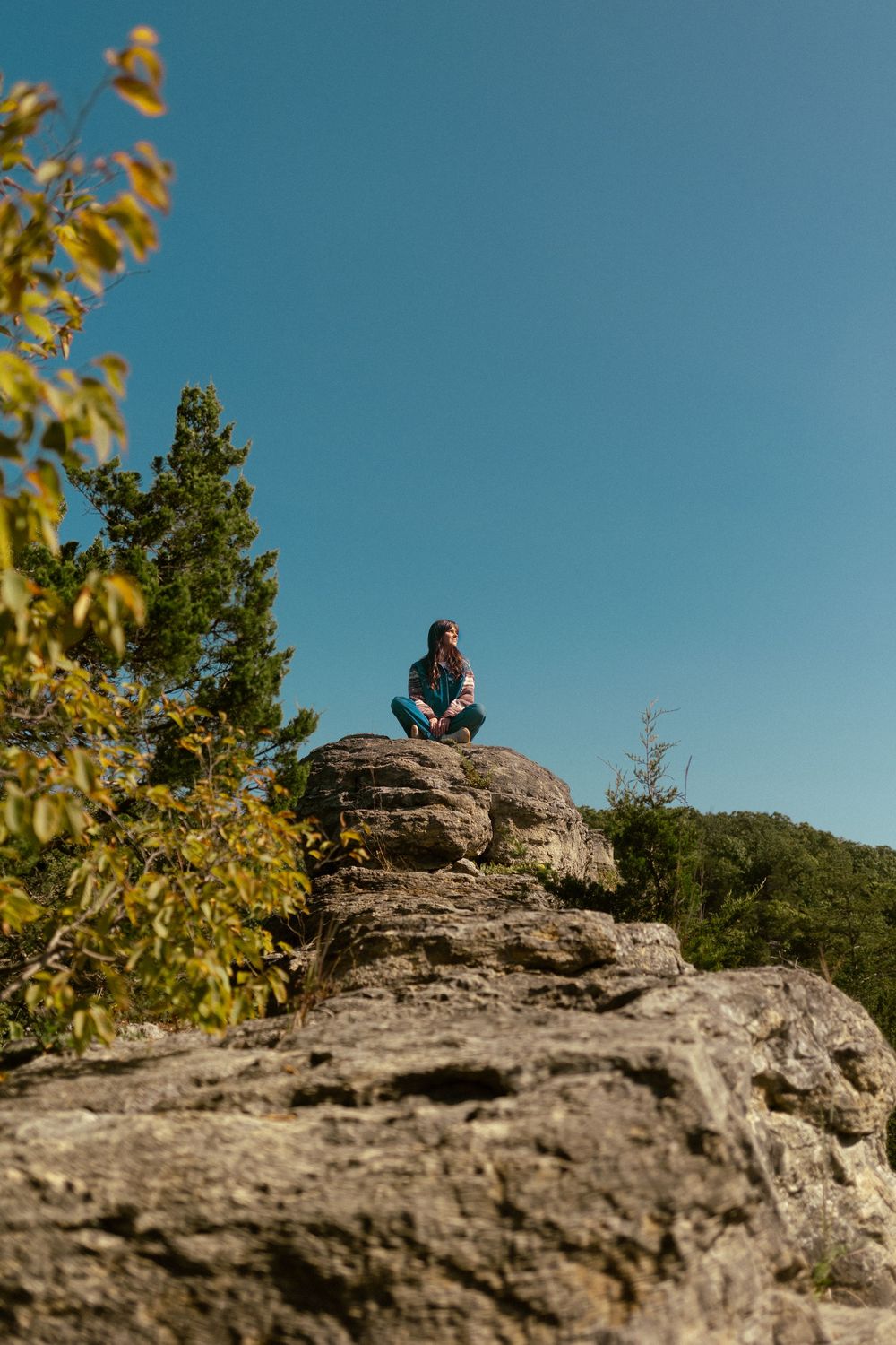 A lone figure sits peacefully on a rocky cliff ledge against a bright blue sky with autumn foliage framing the scene.