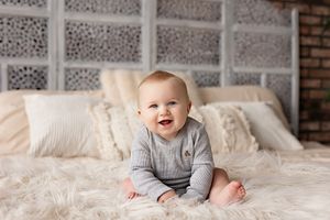 Happy baby in gray sweater sitting on white bedding with textured wall backdrop.