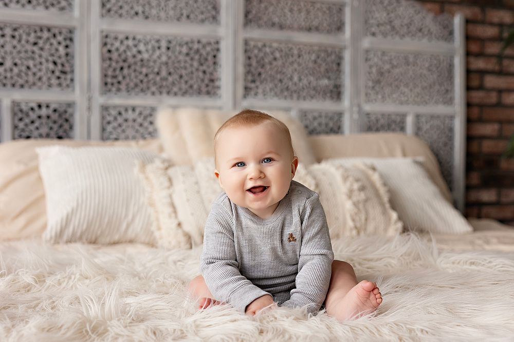 Happy baby in gray sweater sitting on white bedding with textured wall backdrop.