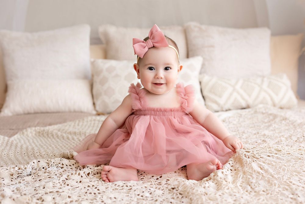 A baby in a pink tutu dress sits on a soft white blanket with delicate lace details and ruffles.  Kendall Kuss Photography Watertown WI