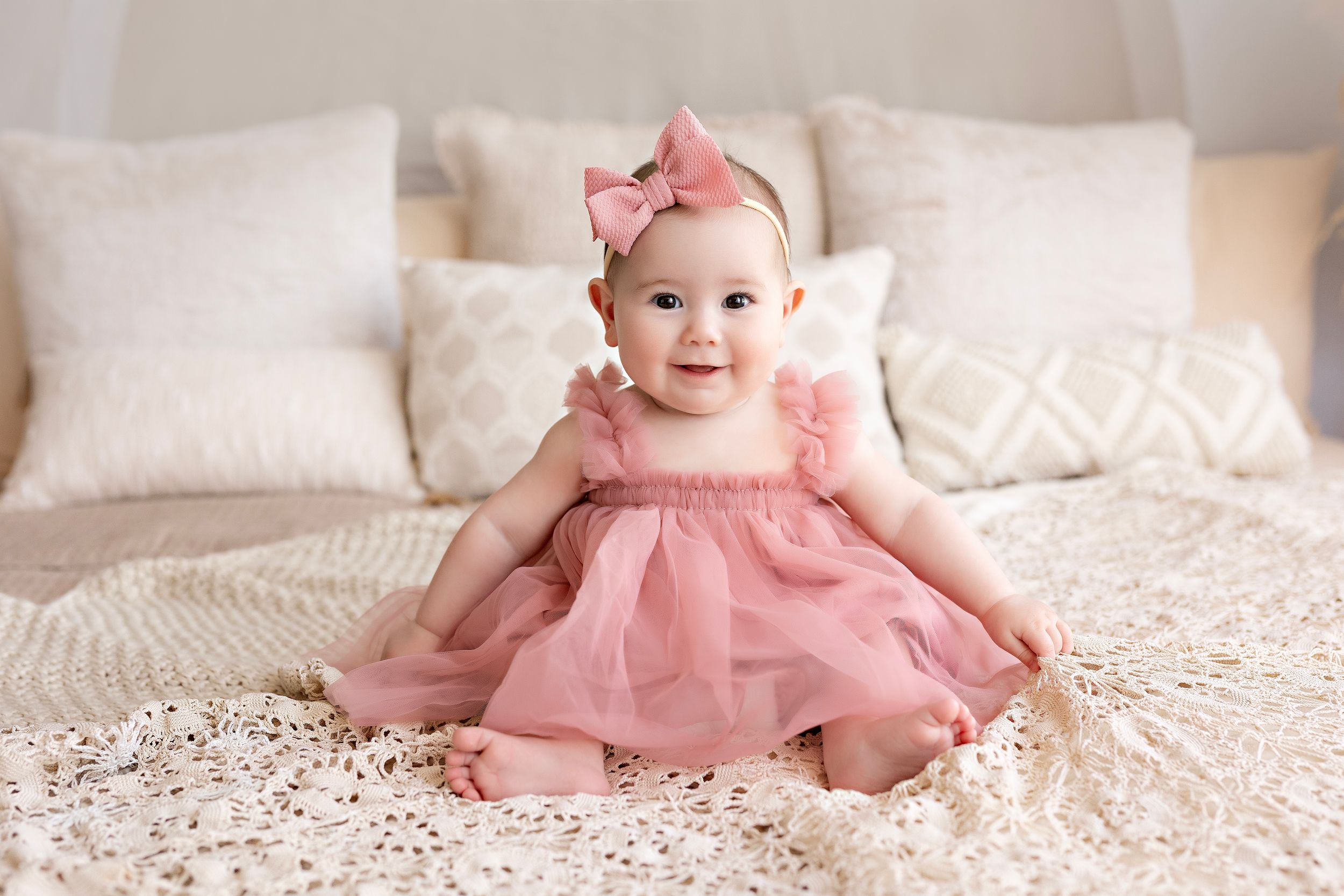 A baby in a pink tutu dress sits on a soft white blanket with delicate lace details and ruffles.  Kendall Kuss Photography Watertown WI