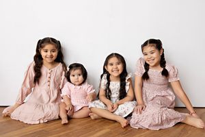 Four children wearing matching pink dresses sit together on a wooden floor against white background.  Kendall Kuss Photography Watertown WI