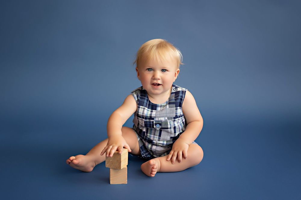 A playful baby in overalls sits and poses against a dark blue backdrop in a photography studio setting.  Kendall Kuss Photography Watertown