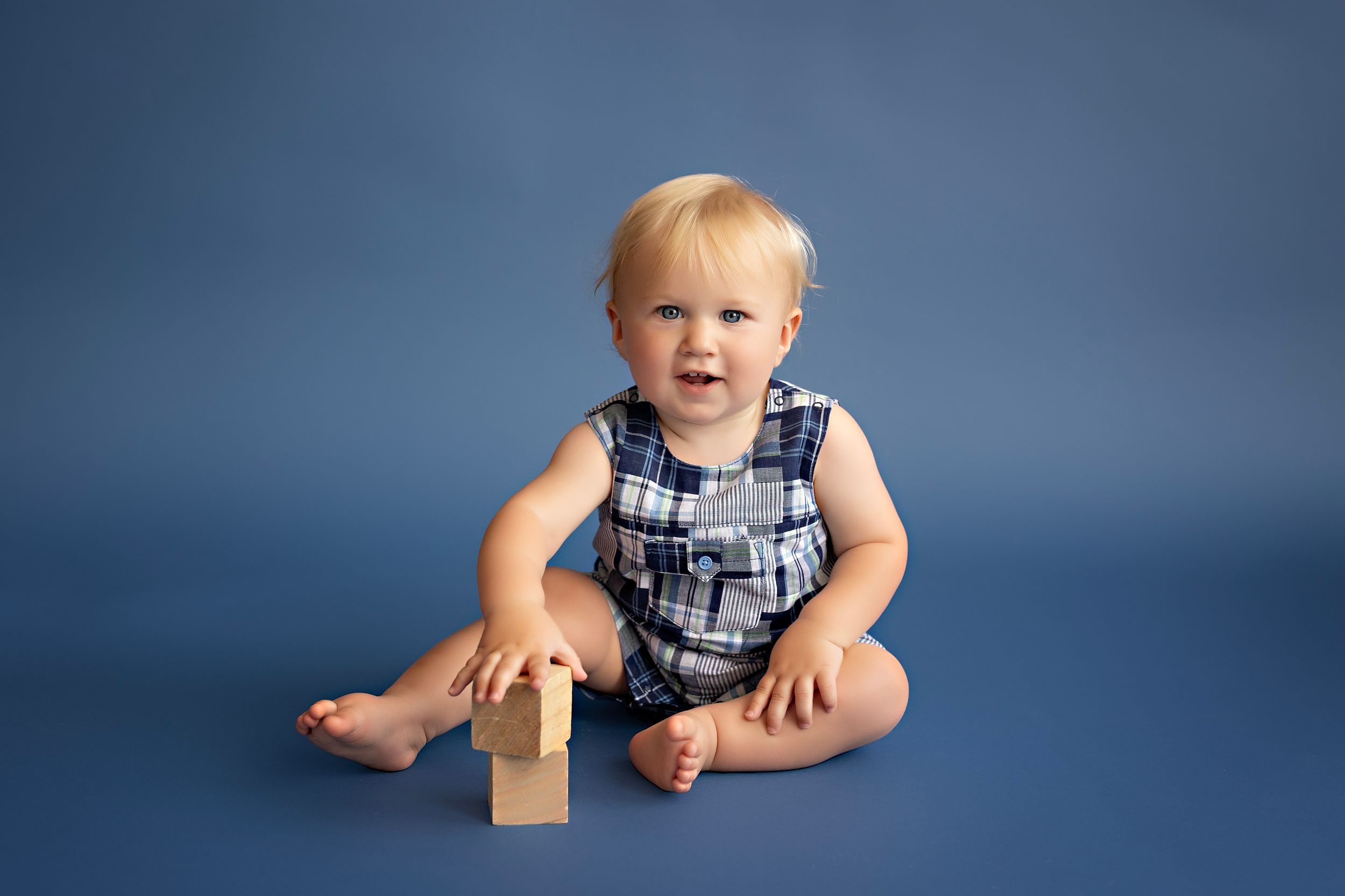 A playful baby in overalls sits and poses against a dark blue backdrop in a photography studio setting.  Kendall Kuss Photography Watertown