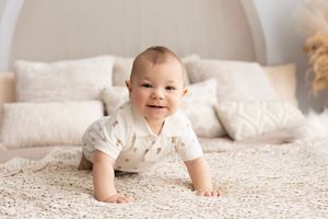 Smiling baby in white outfit crawls on plush cream carpet with soft pillows in background.  Kendall Kuss Photography Watertown WI