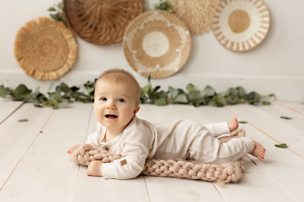 Baby in cream outfit lying on white floor with boho wall decor and greenery. Kendall Kuss Photography baby photos at studio in SE Wisconsin