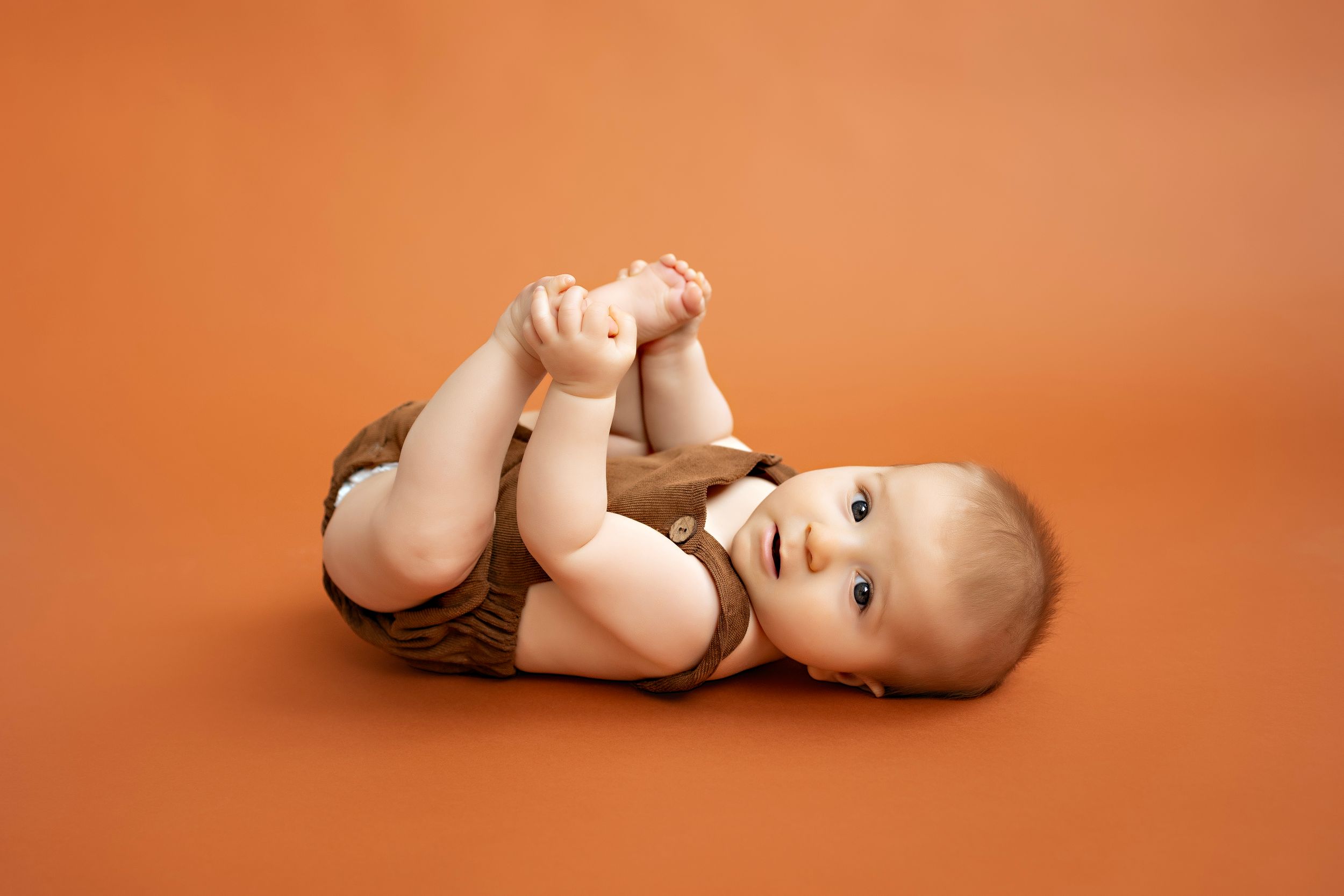 6 month baby boy pose lying on orange backdrop reaching for feet. Kendall Kuss Photography baby photos at studio in Watertown WI