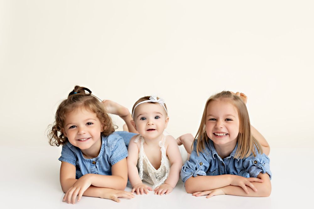 Three siblings posed lying on white background in matching blue outfits during studio photoshoot. Kendall Kuss Photography sibling photos