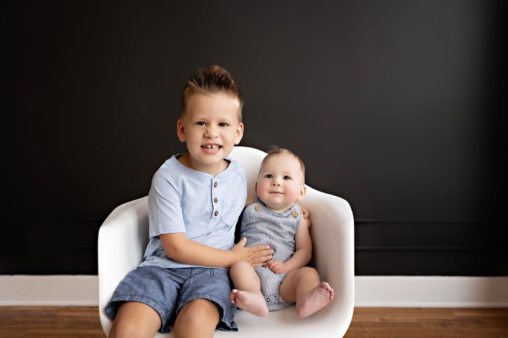 Two siblings sitting together on white chair against dark wall during portrait session. Kendall Kuss Photography baby photos at studio in WI