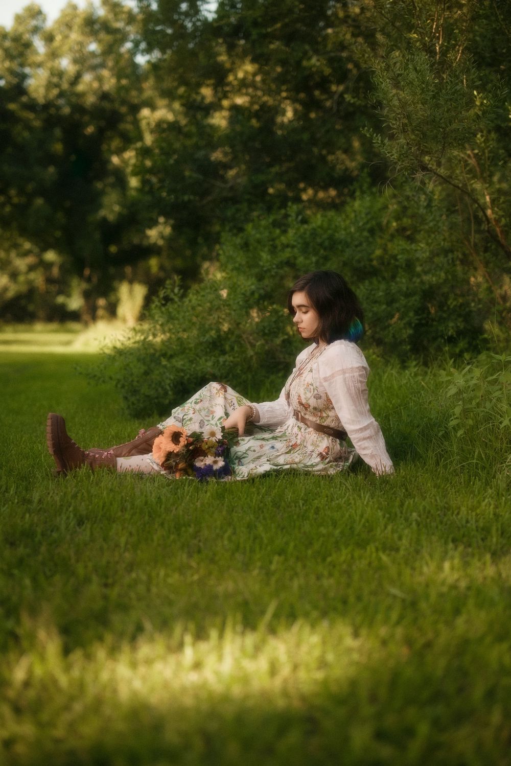 Young woman in floral dress and brown boots sitting on lush green grass holding a bouquet of wildflowers near trees.