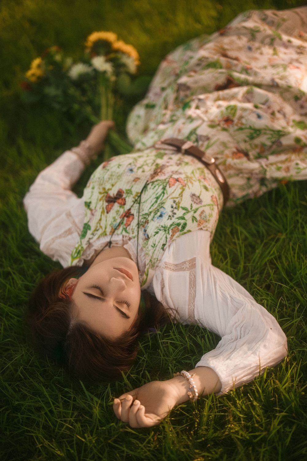 Woman in floral dress lying on green grass with eyes closed, holding wildflowers in warm sunlight.