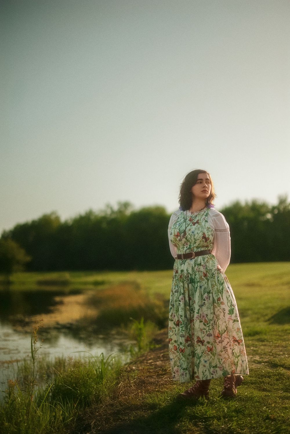 Woman in floral midi dress standing by a peaceful pond in golden hour sunlight with lush green trees behind her.