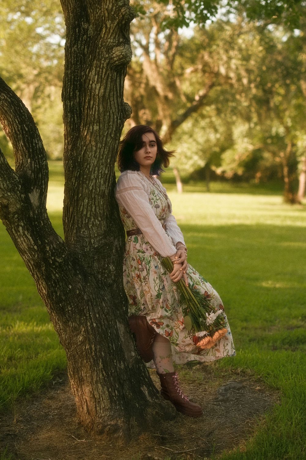 Woman in floral dress leaning against tree in sunlit park, holding wildflowers, wearing brown boots.