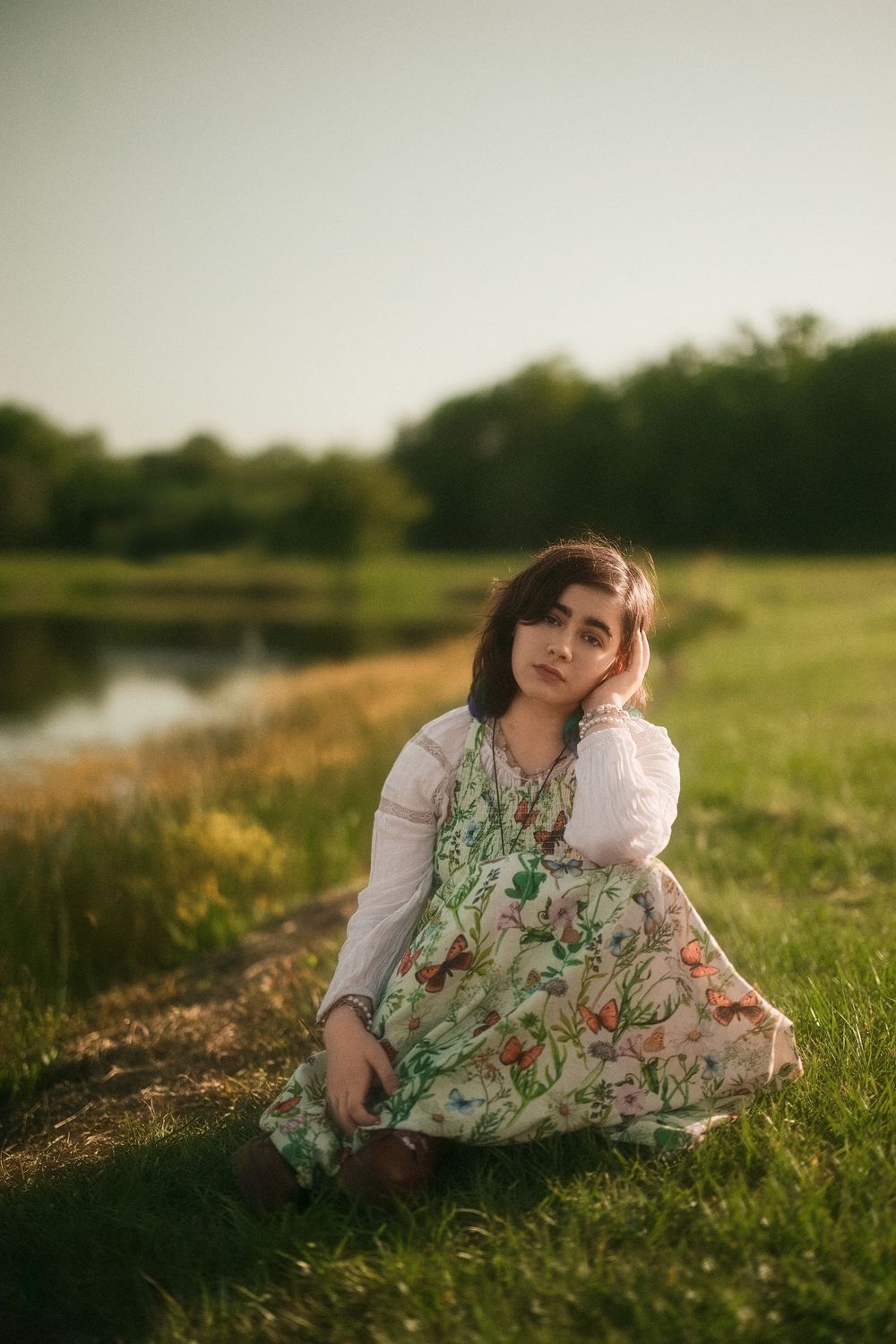 Young woman in floral dress sitting on grass by a pond at golden hour in a serene outdoor setting.