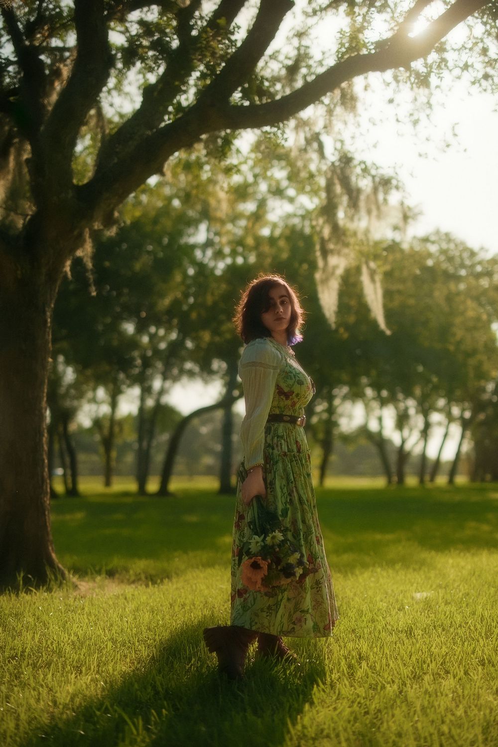 A woman in a floral green dress stands under oak trees in golden sunlight, holding wildflowers in a lush green park.