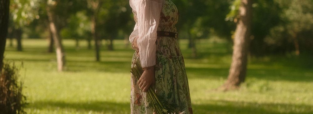 Woman in floral green skirt and pink top walking in a sunny park with lush green grass and trees.