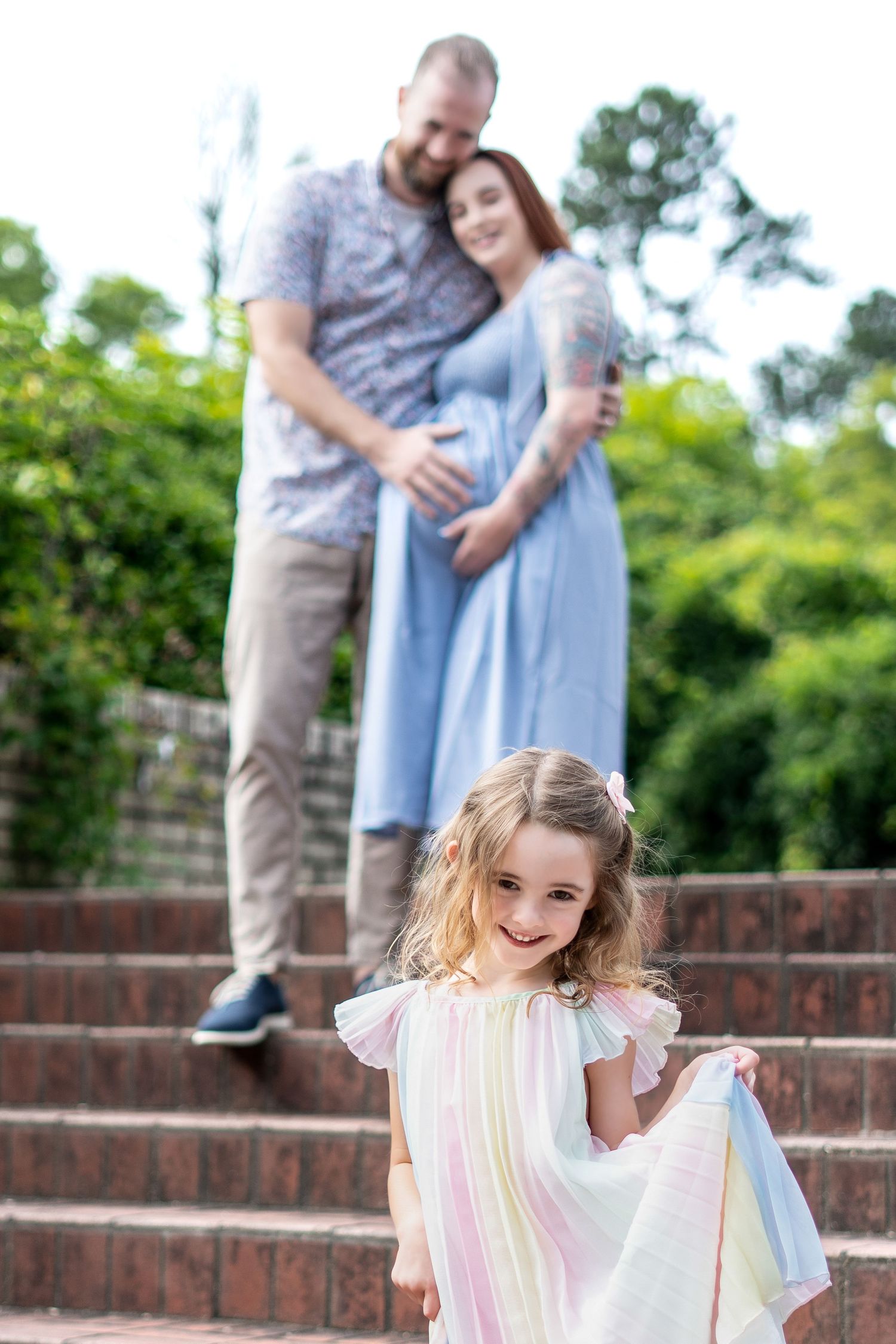 Family portrait on brick steps with young girl in striped dress and expecting parents in background.